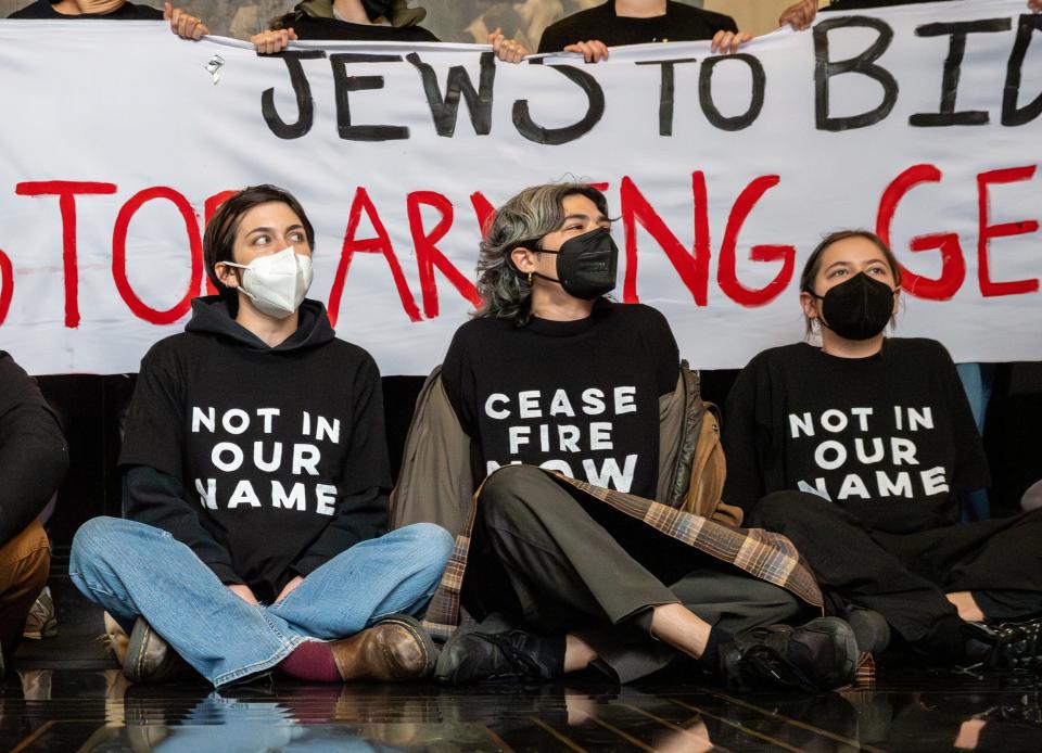 Protesters seated with a banner reading "Jews to BIDEN: STOP AIDING GENOCIDE" and wearing shirts with "CEASE FIRE NOW" and "NOT IN OUR NAME"