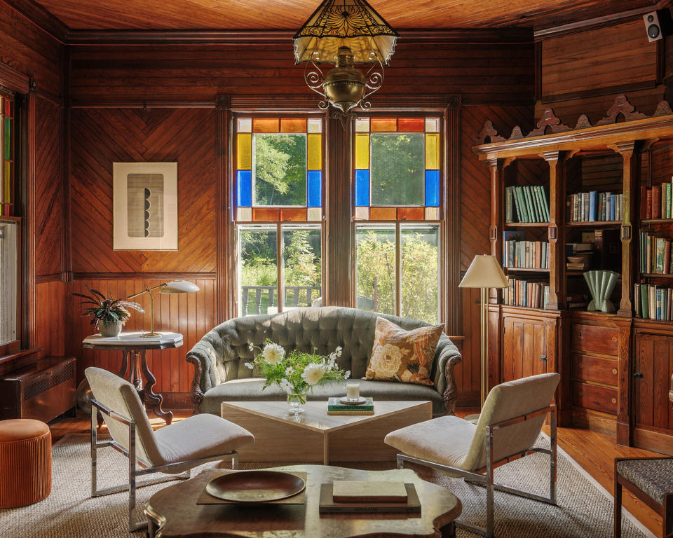 Large living room with dark wooden walls, colorful stain-glass windows, modern lounge chairs and sofa, rug, lamps