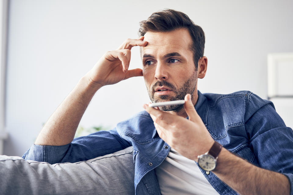 Man in a denim shirt holds phone on speaker, looks away thoughtfully