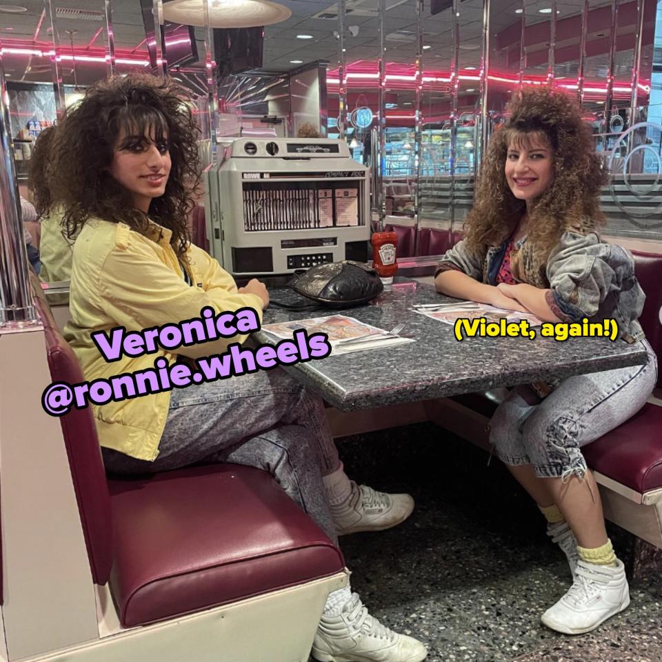 Two women with curly hair and lively 80s fashion sit in a retro diner booth, facing each other with smiles