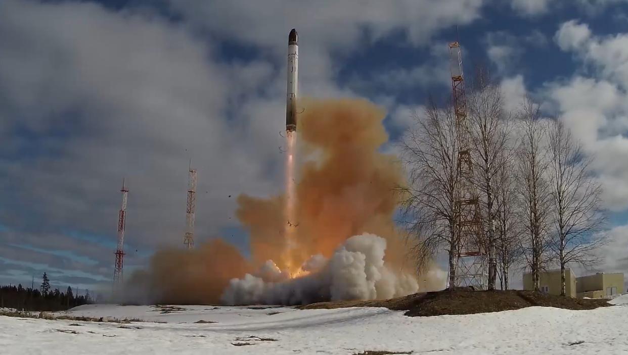 A missile ejects flames and smoke as it takes off from snow-covered ground near three towers, a few buildings and a patch of trees beneath a blue sky dotted with clouds.