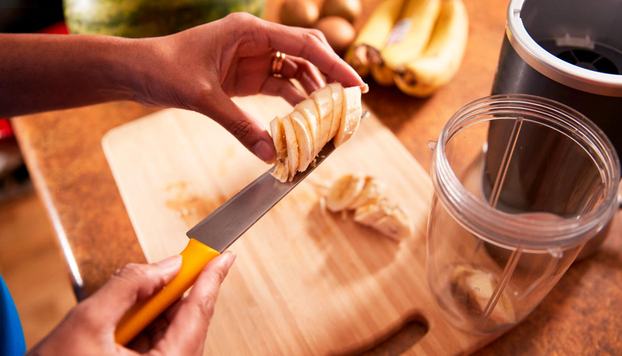  A woman's hands holding a knife with slices of banana on the blade by a smoothie jug. 