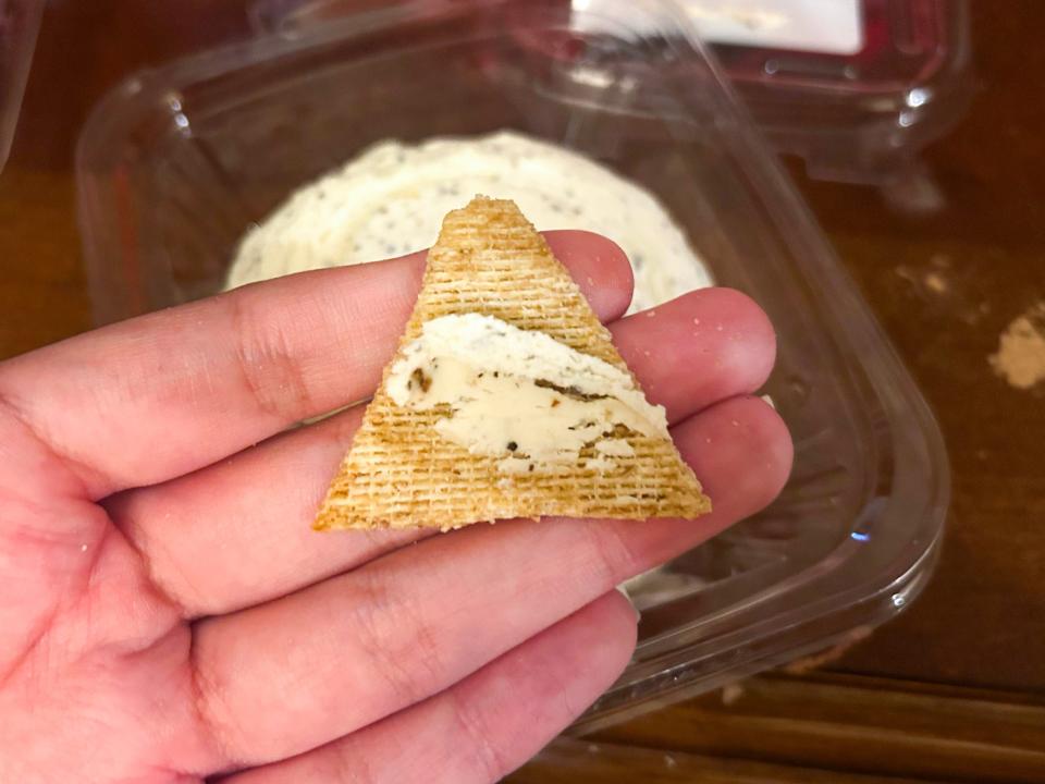A hand holds a triangular cracker with a small scoop of white butter with black specs in it in front of a container of white butter