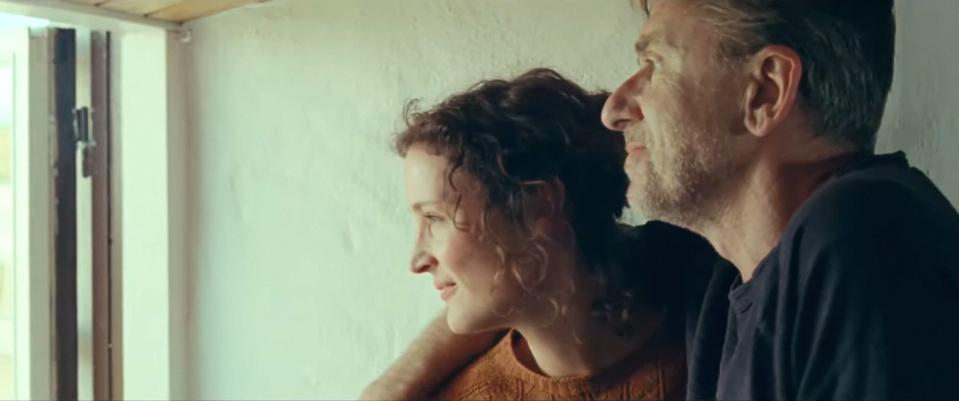 A white man and woman couple stare happily out a window. He has his arm around her.