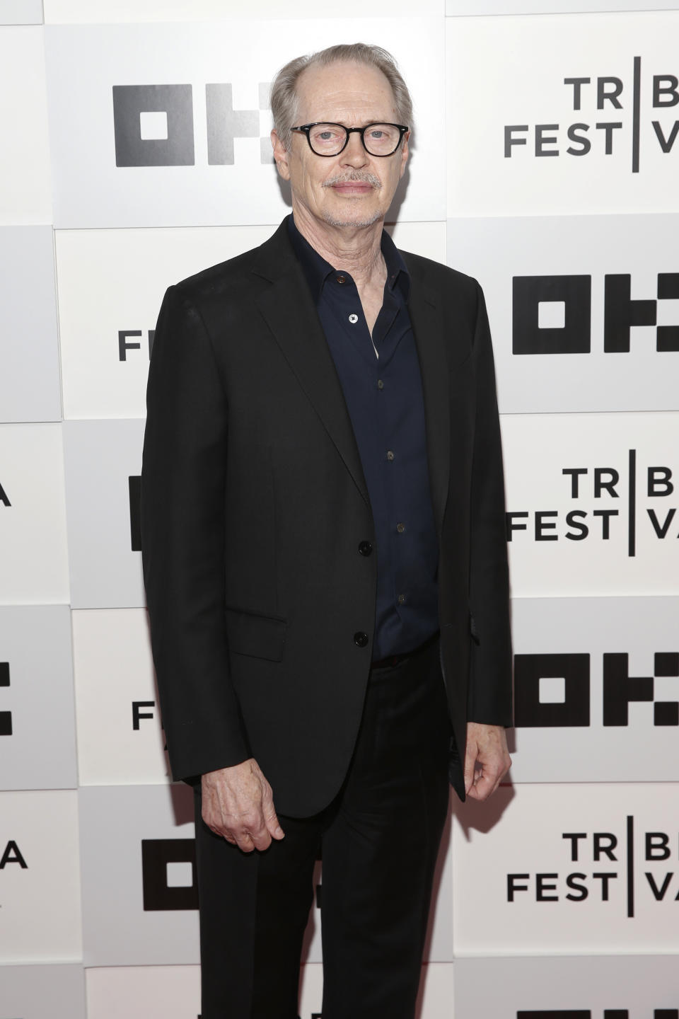 Steve Buscemi at the Tribeca Film Festival, wearing a black suit and dark shirt, standing in front of a festival backdrop