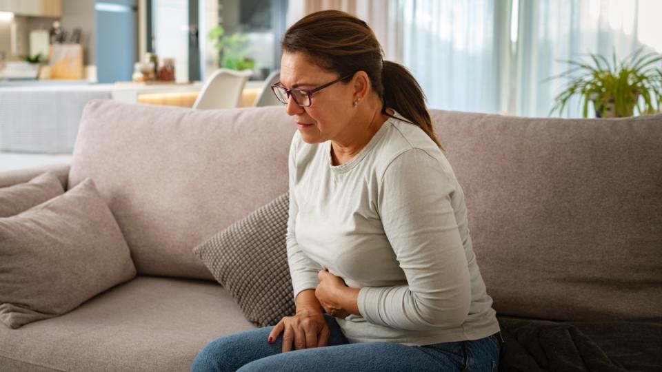 A brunette woman sitting on a couch while clutching a hand to her stomach, in pain due to one of two stomach ulcer causes