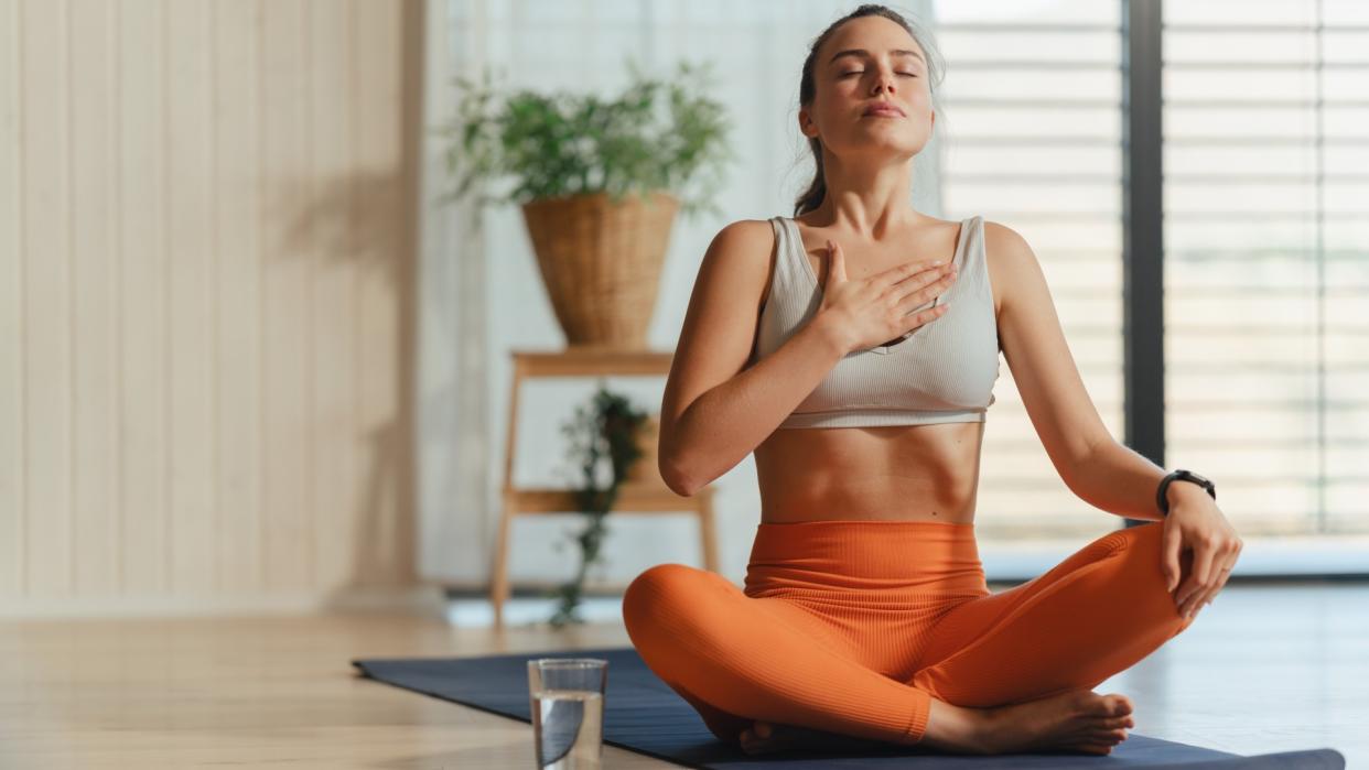  Woman sitting on yoga mat in light and airy studio cross-legged performing a breathing exercise with hand on chest. 