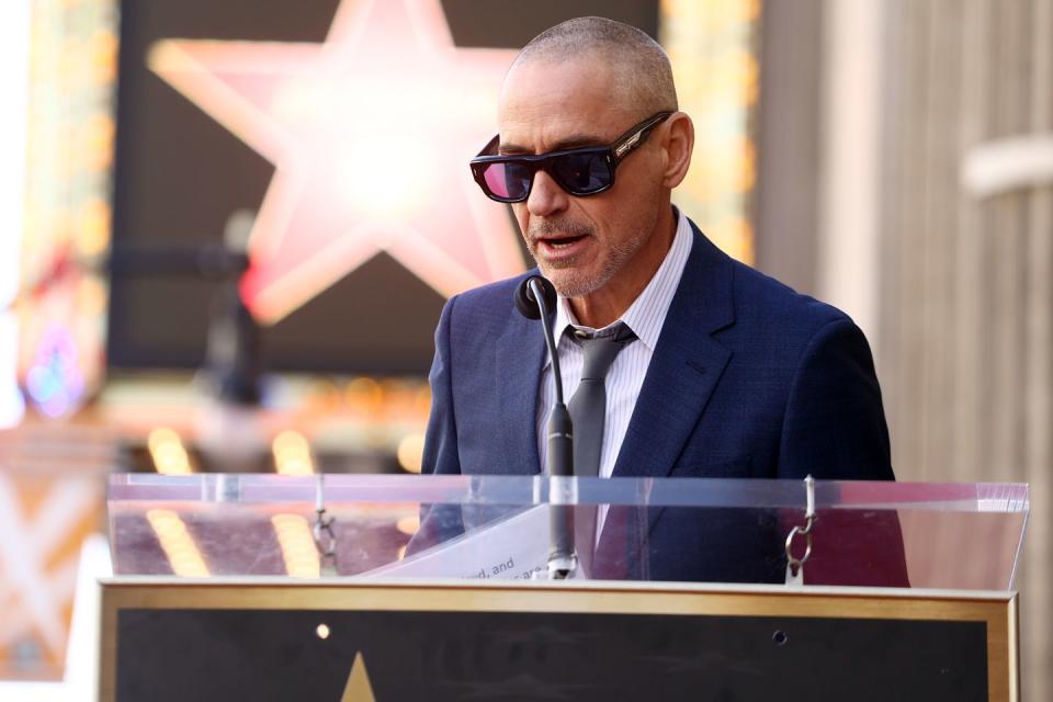 robert downey jr speaks into a microphone while wearing sunglasses and a blue suit jacket over a white striped shirt and gray tie