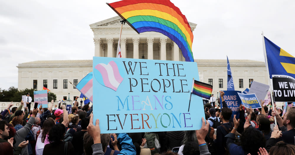 The HRC has declared the a state of emergency for LGBTQ+ people in the US. The image shows a large group demonstrating. They are flying rainbow flags with a placard which reads 