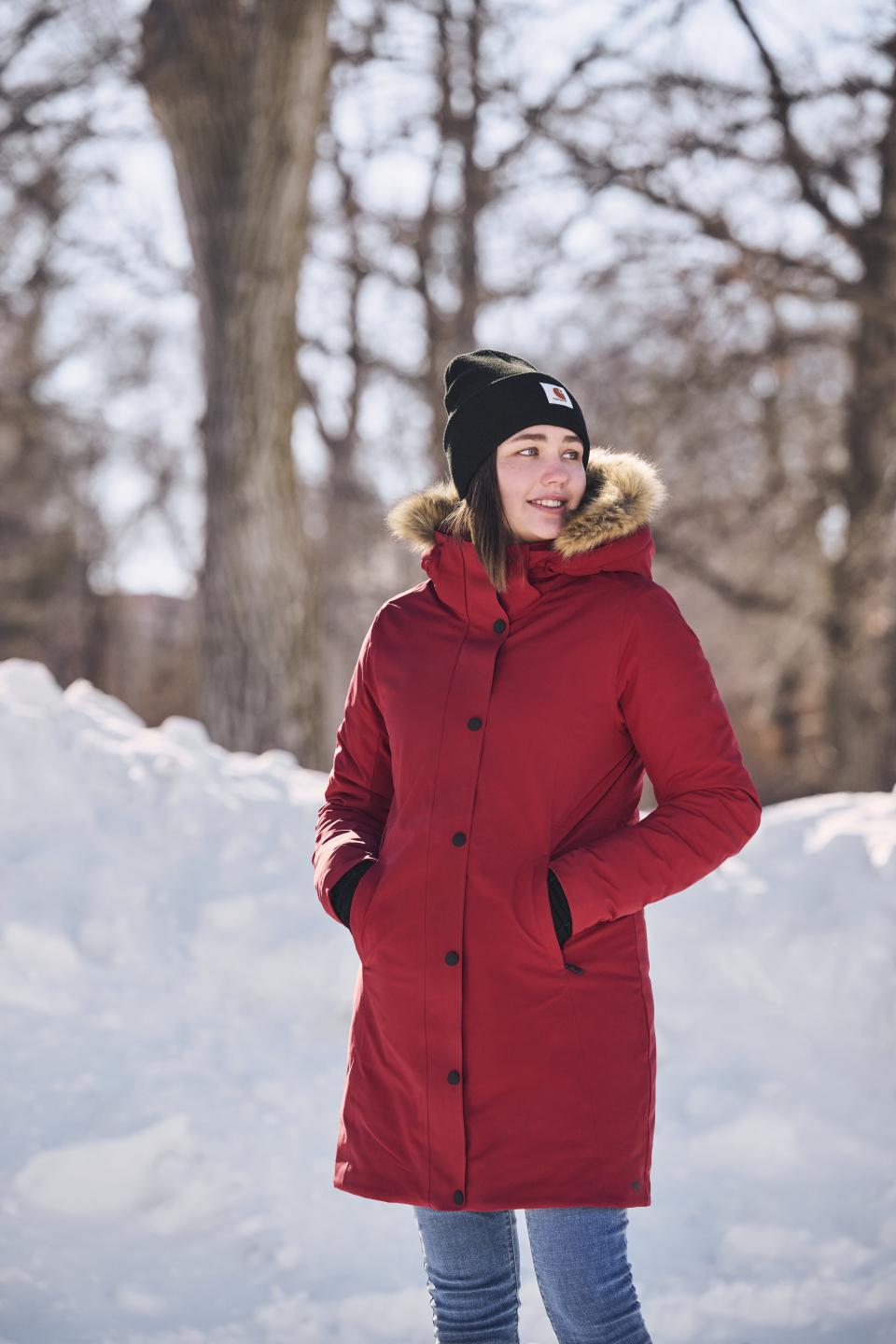 A woman in a winter parka and toque from Mark's stands next to a snowbank looking off in the distance