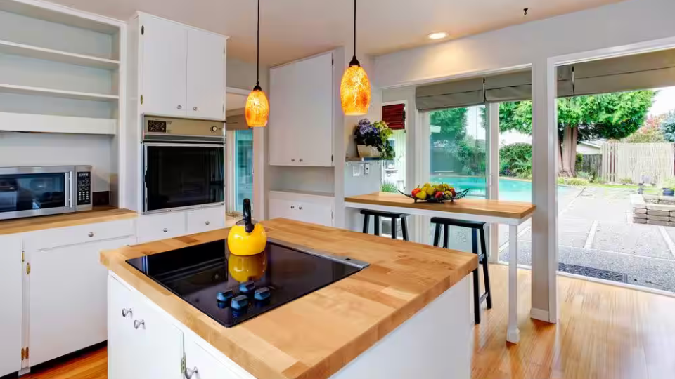 A white indoor kitchen with butcher block countertops is lit by warm lights and sunlight from open shutter doors.