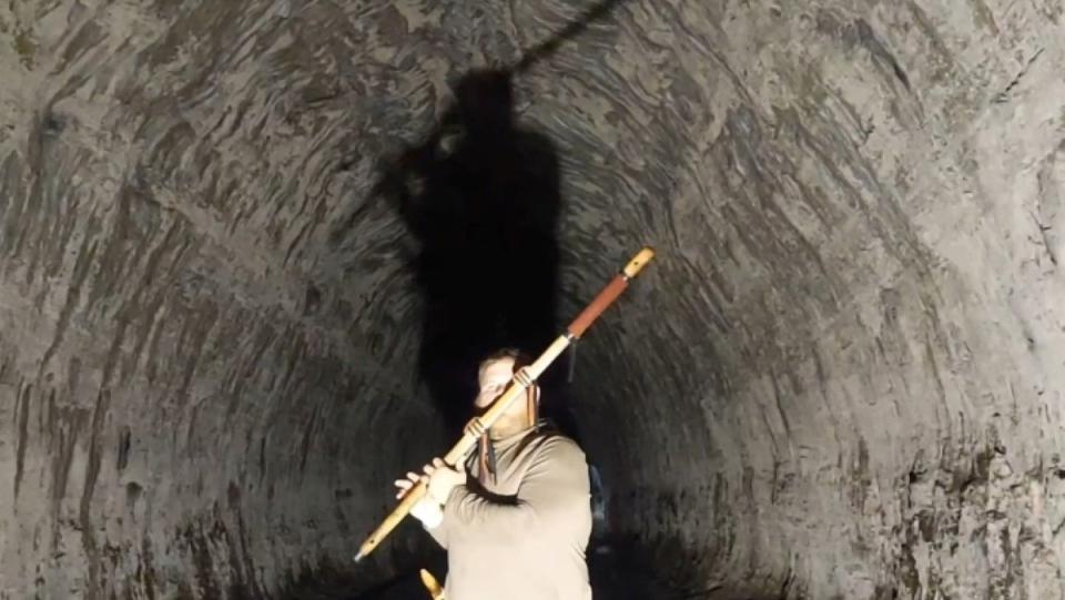 A man playing a long wooden flute inside a cave