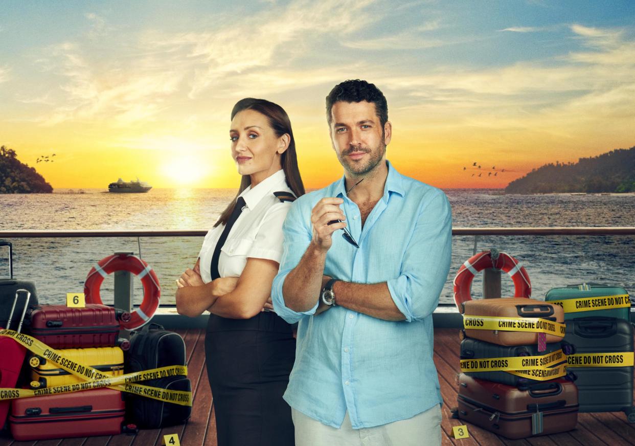  First Officer Kate Woods (Catherine Tyldesley) and Jack Grayling (Shayne Ward) stand on the deck of a ship with a beautiful Mediterranean sunset behind them. On the deck behind them are lots of suitcases wrapped in neon yellow tape marked 'crime scene do not cross'. 