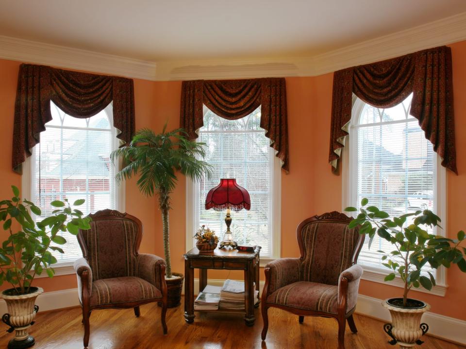 Living room with two chairs arranged around a table next to potted plants and orange walls. Deep red window treatments frame three windows.
