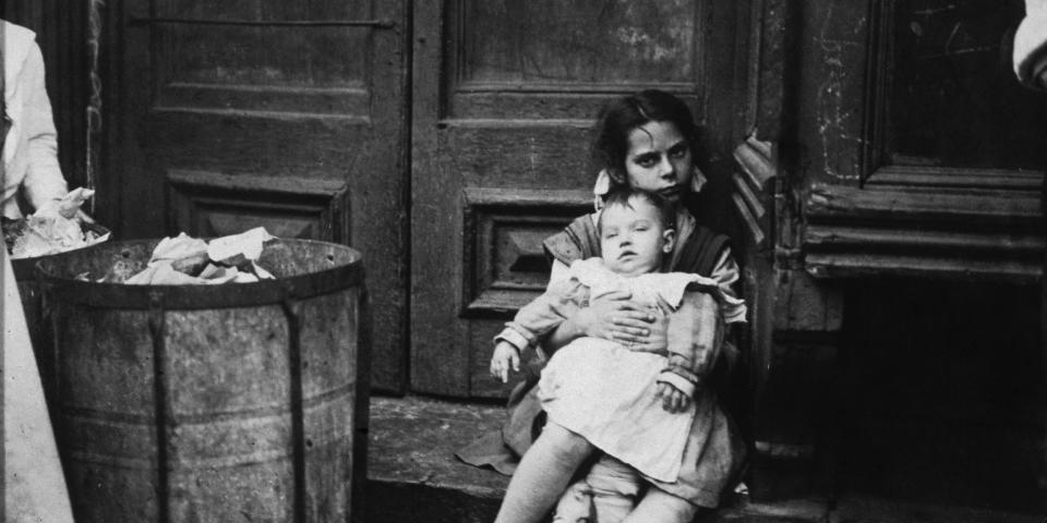 A young girl holding a baby sits in a doorway in New York City, circa 1890.