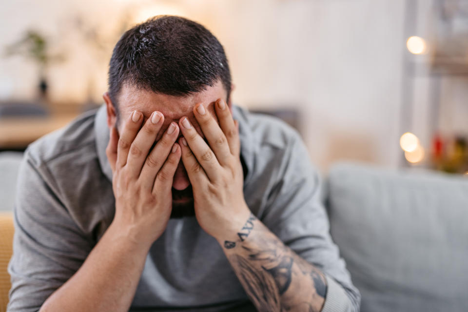 A person with a beard and tattoos is sitting on a couch, covering their face with their hands in apparent distress