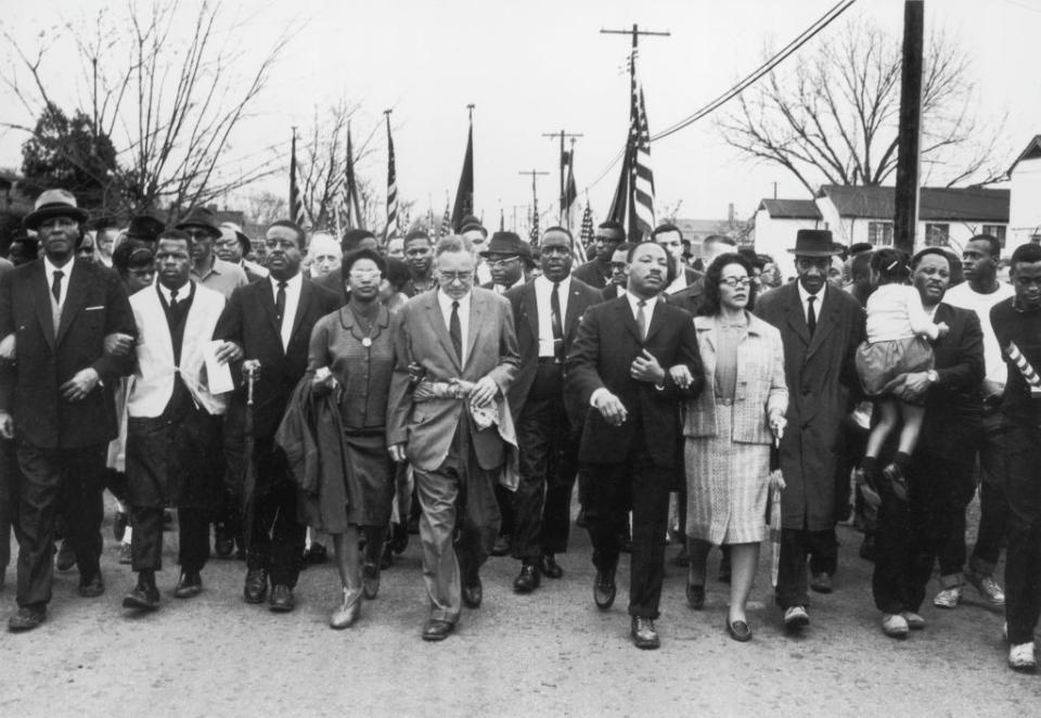 a group of many people including martin luther king jr and coretta scott king walk arm in arm on a city stree, houses and the tops of american flags can been seen in the background