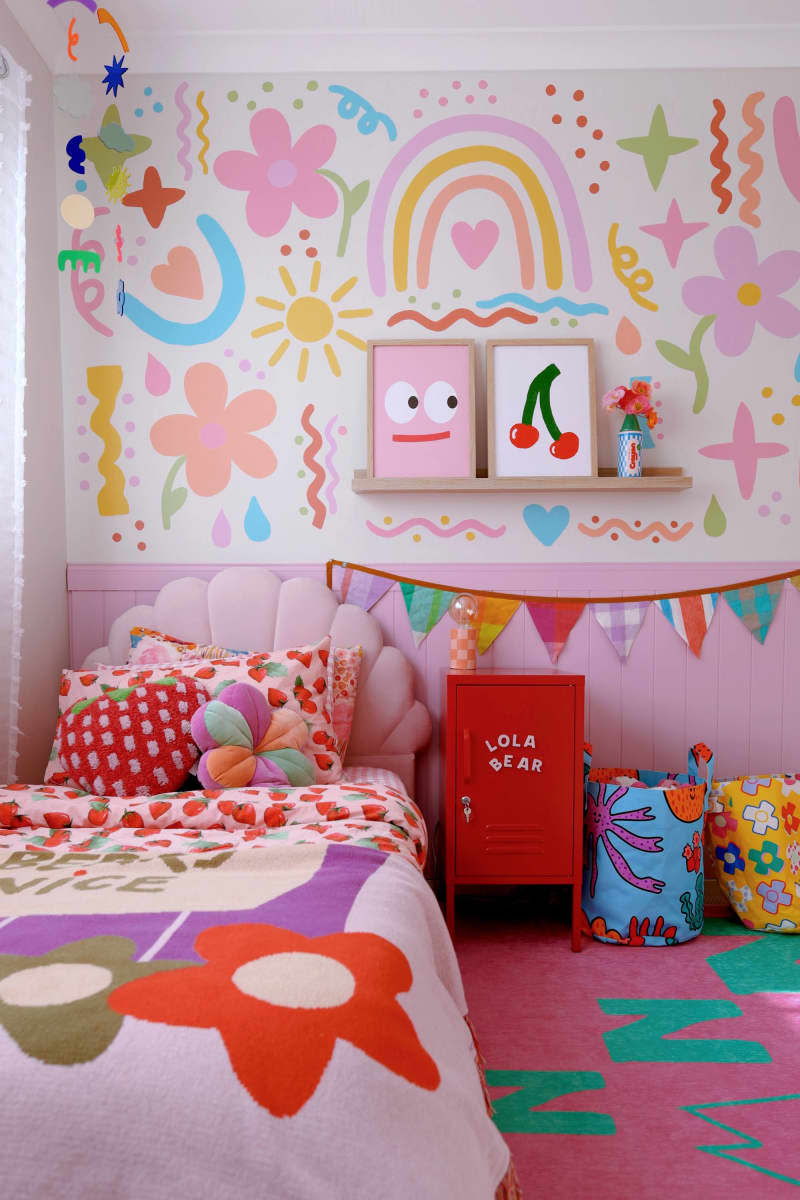 Rainbow squiggles and doodle mural in pinks, yellows, green, and blue, pink paneling half wall, pink and teal zzz carpet, large flowers on bed spread, floating shelf for cherry art