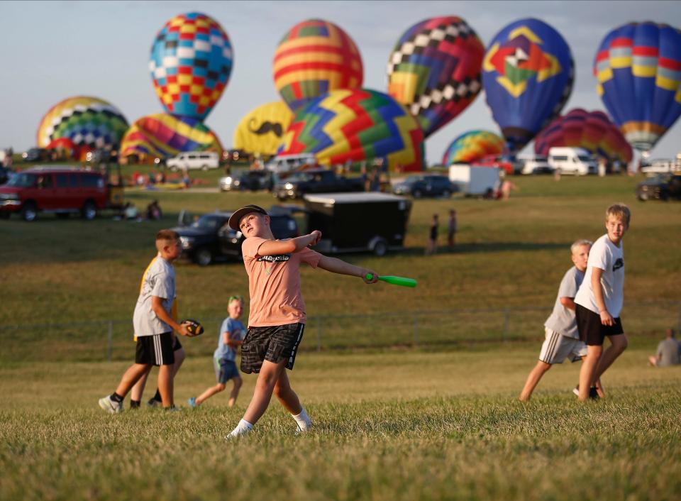 Madden Kiester, 10, of Altoona, hits a ball while playing baseball as the hot air balloons inflate in the background during the National Balloon Classic in 2022.