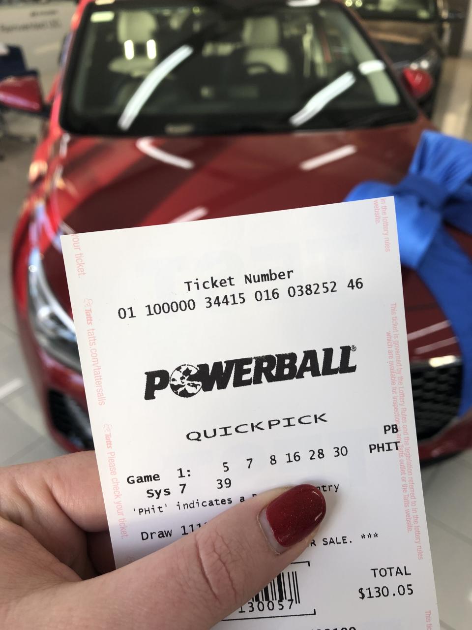 A woman holds a Powerball ticket in front of a red car. 