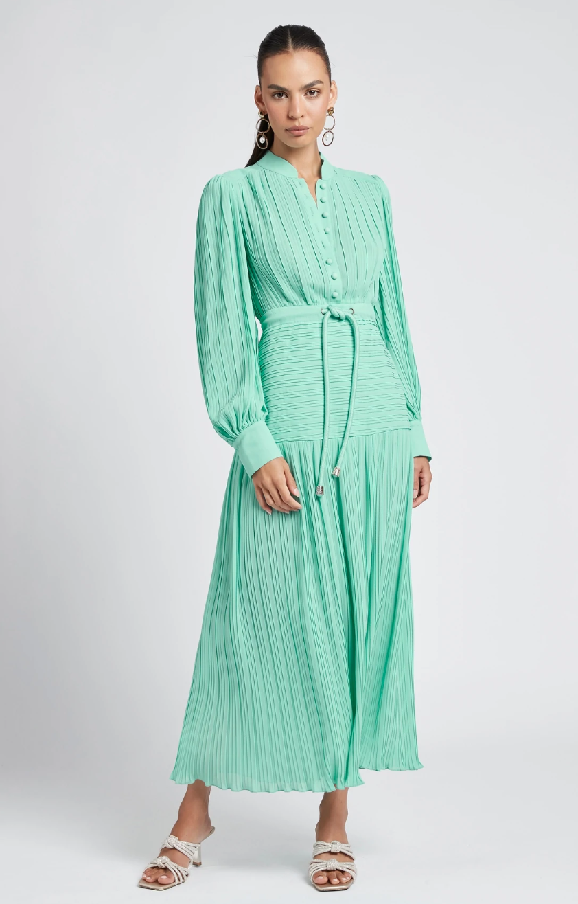 A model wears a mint green maxi dress with long puffed sleeves, waist tie, against a grey background.