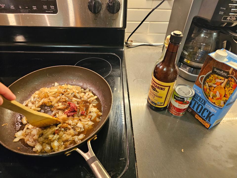 Onions in brown liquid in pan on stovetop next to tomato paste, Worcestire sauce, and chicken stock on counter