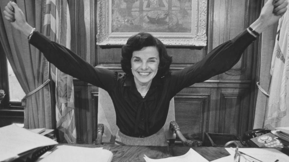 a black and white photo of dianne feinstein stretching her arms in victory and smiling as she sits at a desk in an office