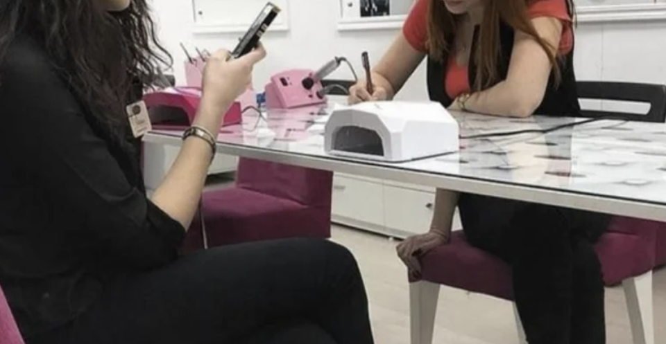 Two women seated at a nail salon table. One checks her phone while the other writes on a notepad. aangesloten apparaten pay for manicures?