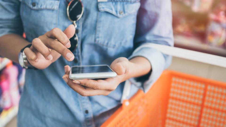 Woman wearing blue jeans shirt and sunglasses using mobile phone to compare price and holding orange shopping basket in mini mart background.