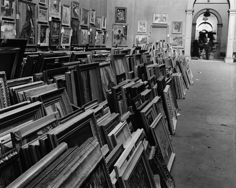 Rows of framed artworks leaning against a wall in a gallery with people in the distance