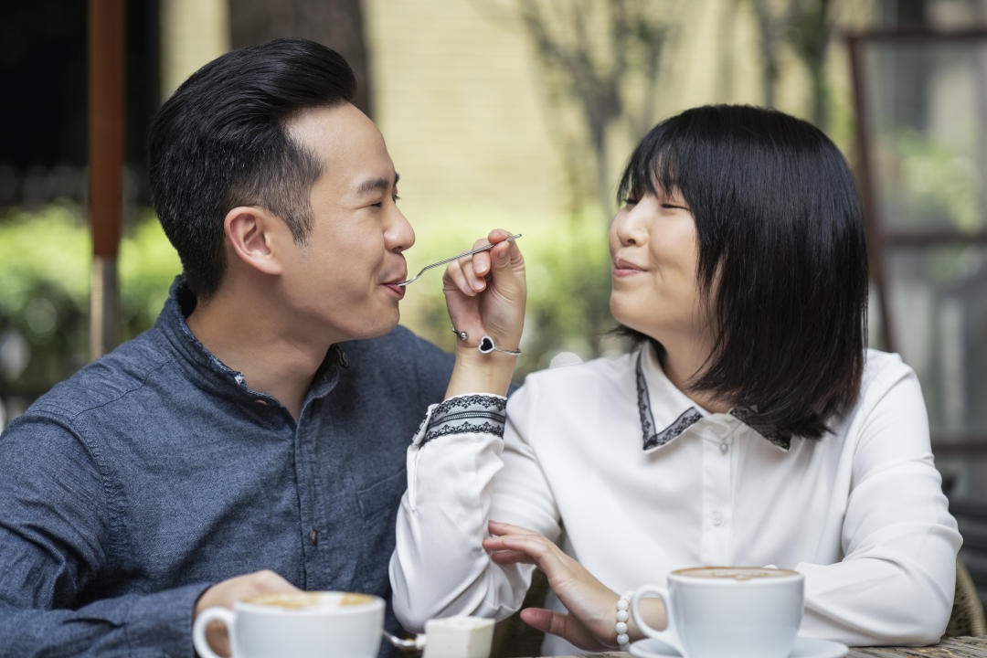 Beautiful romantic woman feeding man with fork at coffee shop. Happy couple is bonding while sitting at table in cafe. They are spending leisure time during weekend.