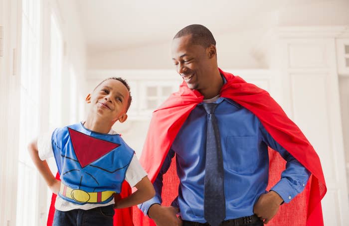 A man and a young boy, both wearing red capes, are smiling at each other. The boy is also in a superhero costume with a belt and emblem