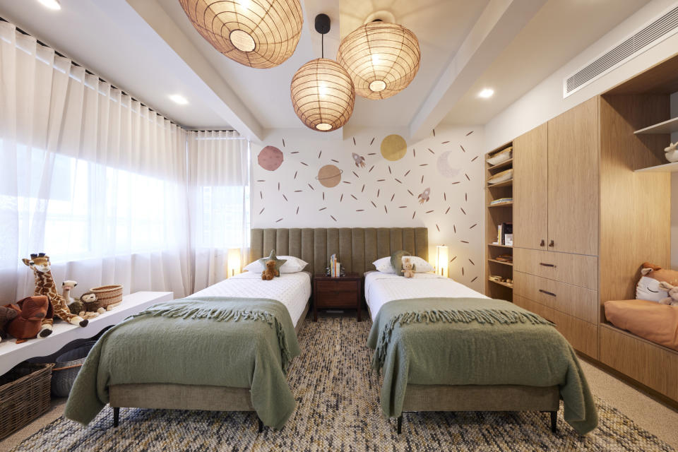 A wide shot of the bedroom with two single beds side by side, the same material light fixtures and rug, a wall of cupboards on the right and window on the left. 