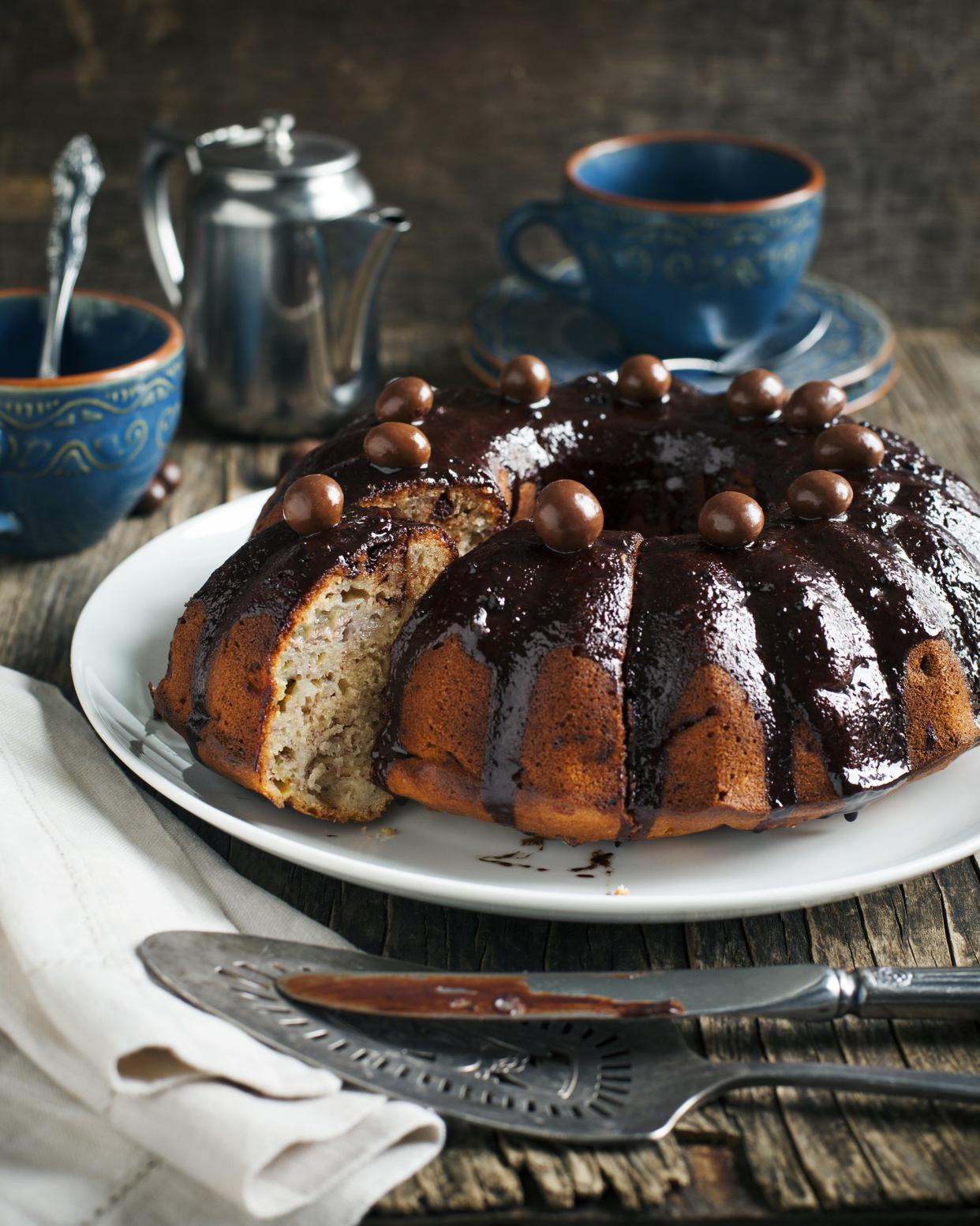 Cinnamon banana cake with chocolate ganacheon a white plate with serving utensils and a white napkin on a dark wooden, rustic table with old world tea cups and a server in the background