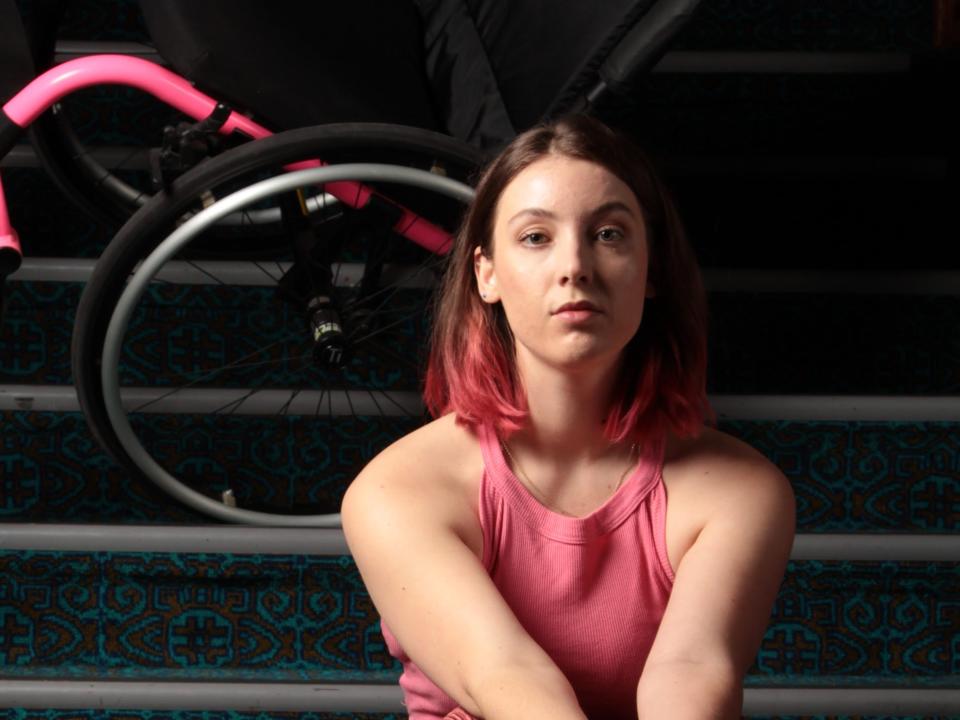 Photograph of Jamila Main sat on stairs with her black and pink wheelchair behind her.