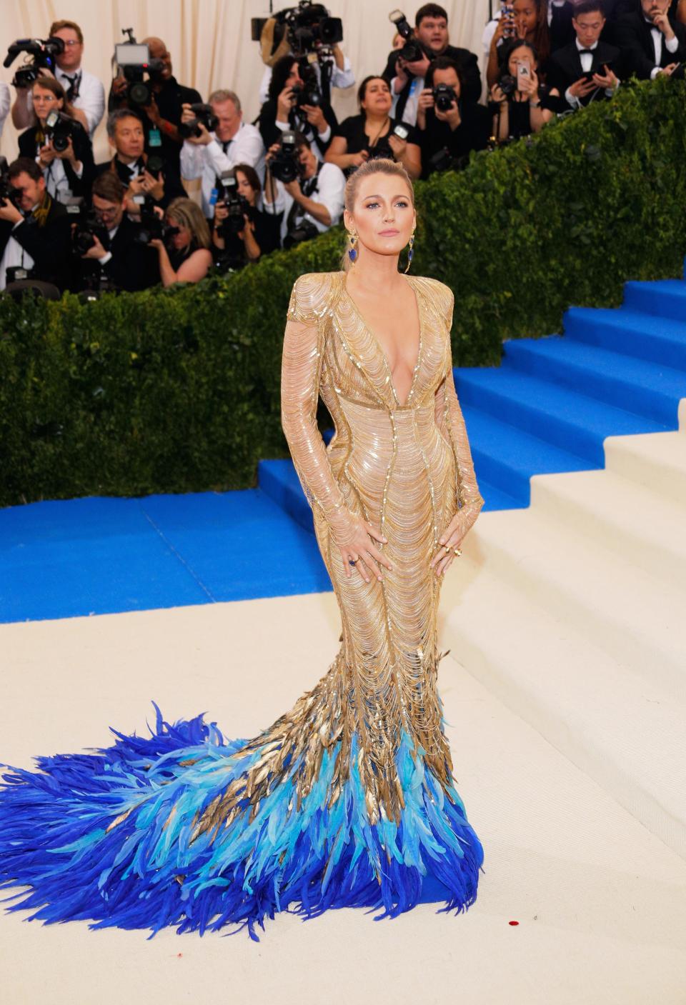 Blake Lively at the 2017 Met Gala in a gold gown with a plunging neckline and blue feathers at the bottom of the skirt.