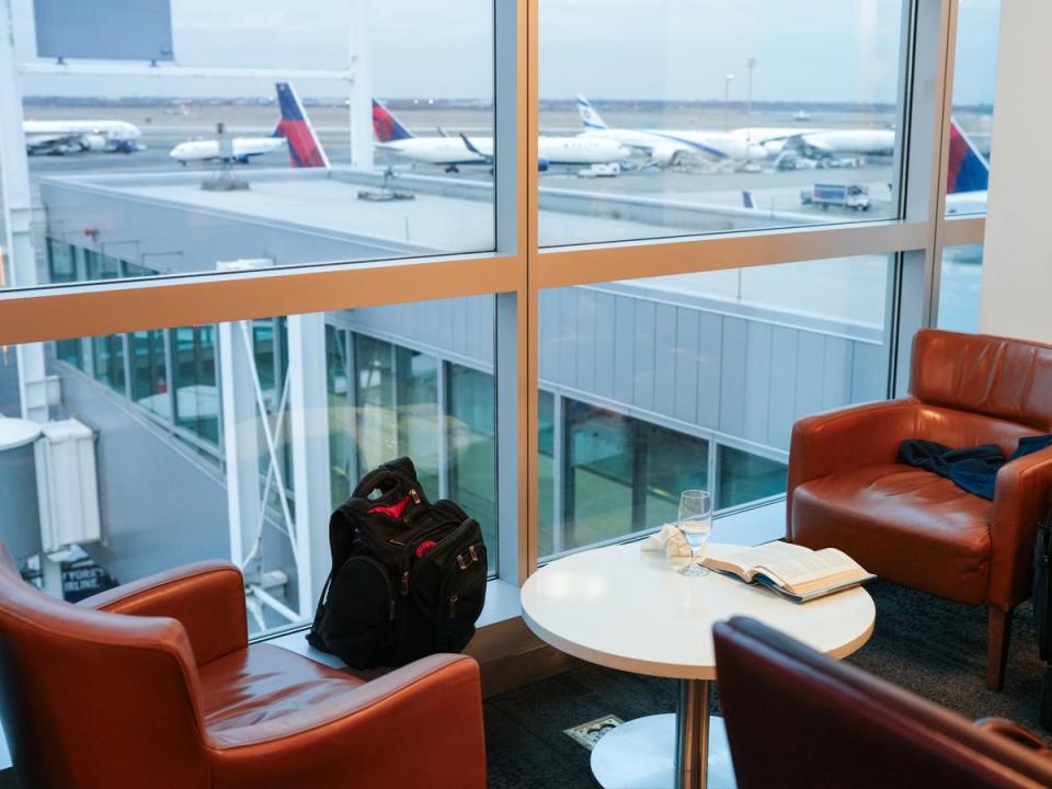 Three red chairs surrounding a table with a book and a glass on it in front of a window with airplanes on the runway