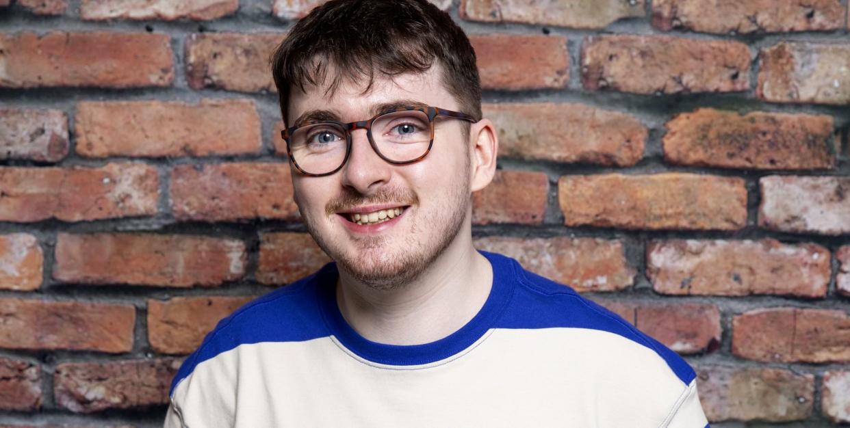 jack carroll as bobby in coronation street, a young man stands with a walking aid and smiles, he wears glasses and a blue and white striped top