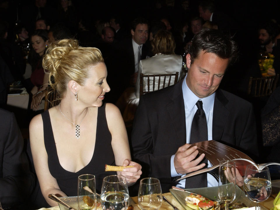 Lisa Kudrow and Matthew Perry sit at a formal event, with Lisa holding food and Matthew looking at a menu or program. Both are dressed in formal attire