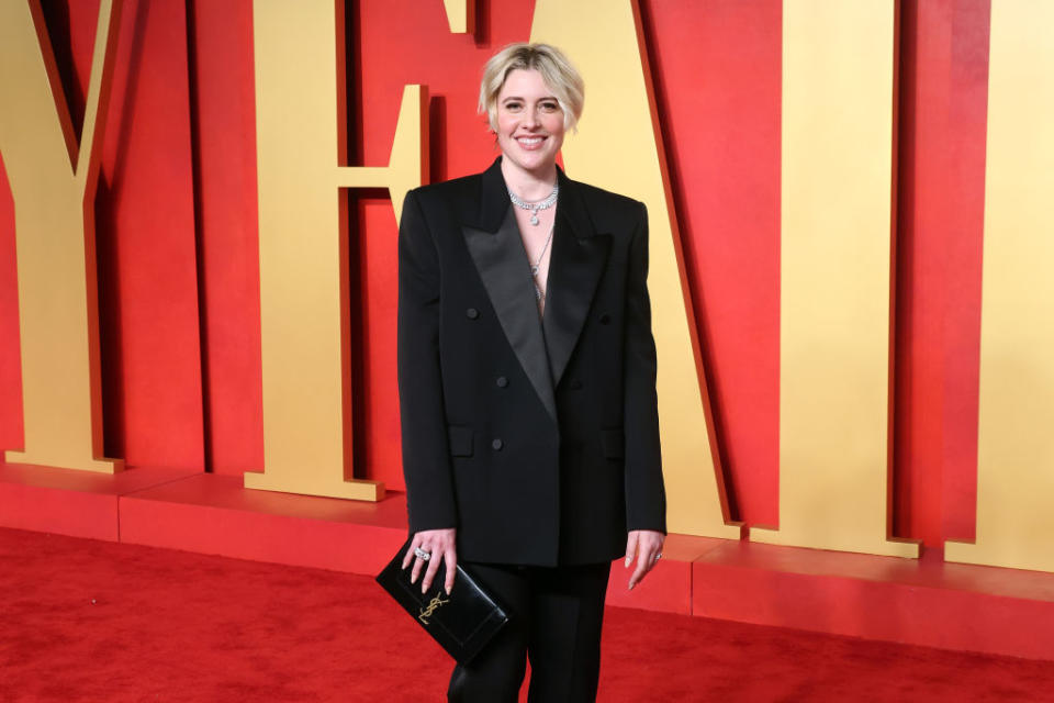 Person in a black suit with a blazer and pants stands smiling on the red carpet