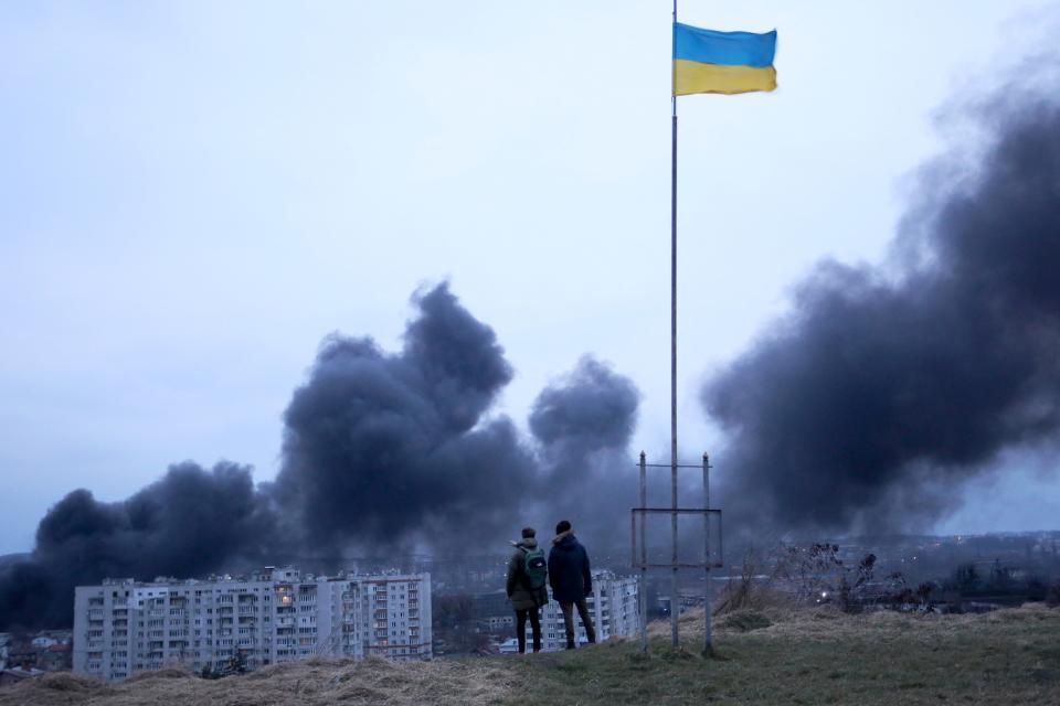 People standing near a Ukrainian national flag watch as dark smoke billows following an airstrike in the western Ukrainian city of Lviv last year. 