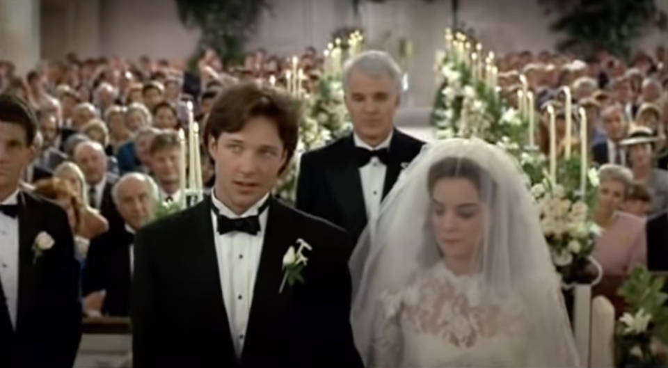 A shot of a bride and groom standing at the altar with the church at their backs, she has a veil over her face and he's in a tux. Steve Martin, also in a tux, is standing behind them
