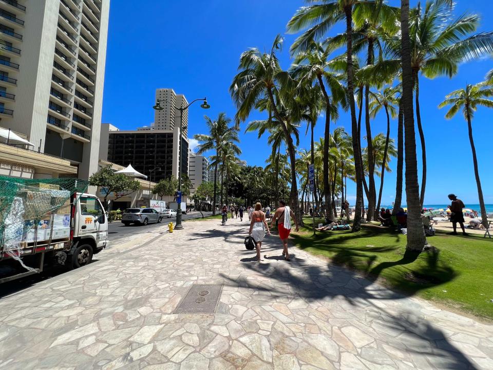 People walking on a path with a green area and beach on one side and buildings and cars on the other.