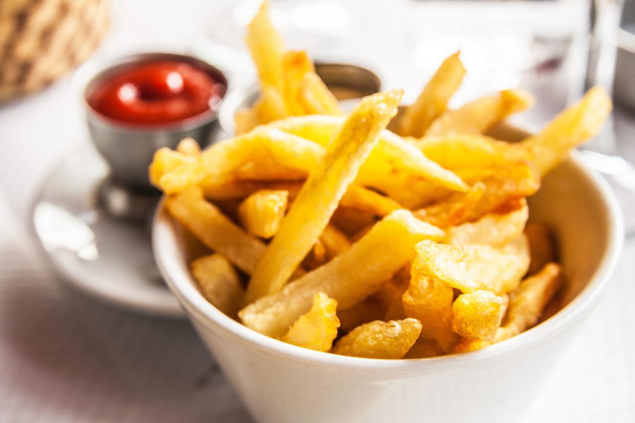 A white bowl of perfect french fries with a blurred background of condiments