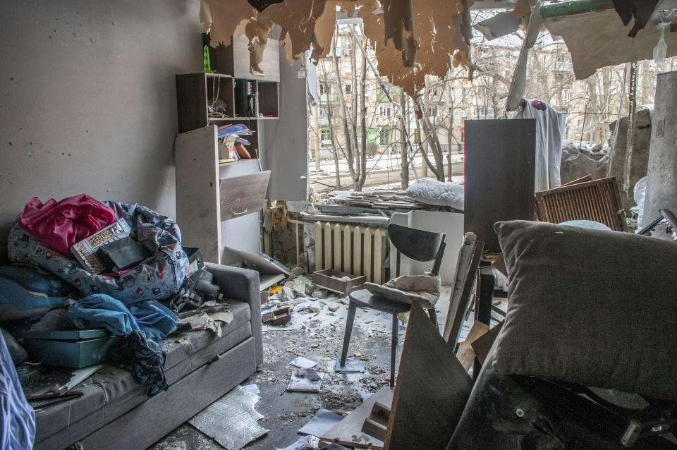 The wreckage of an apartment damaged by shelling, with clothes piled on a couch and streamers of wallpaper dangling..