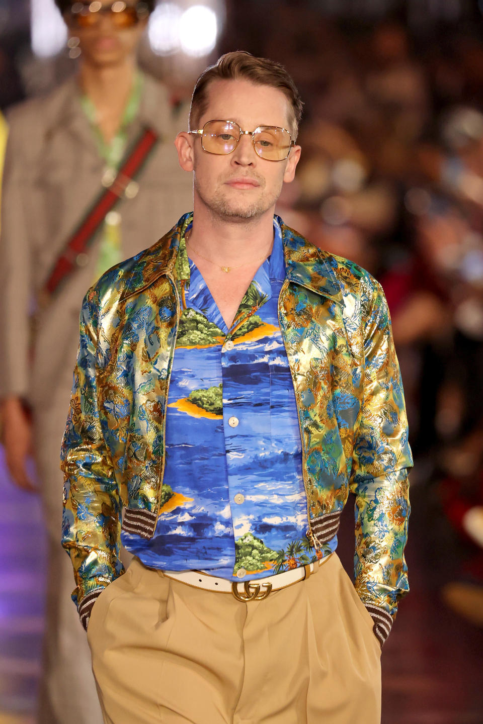 Macaulay on Prada runway, wearing a bomber jacket in the colours of a peacock's tail over a blue shirt with the sea and several small islands on it, and camel colored trousers.