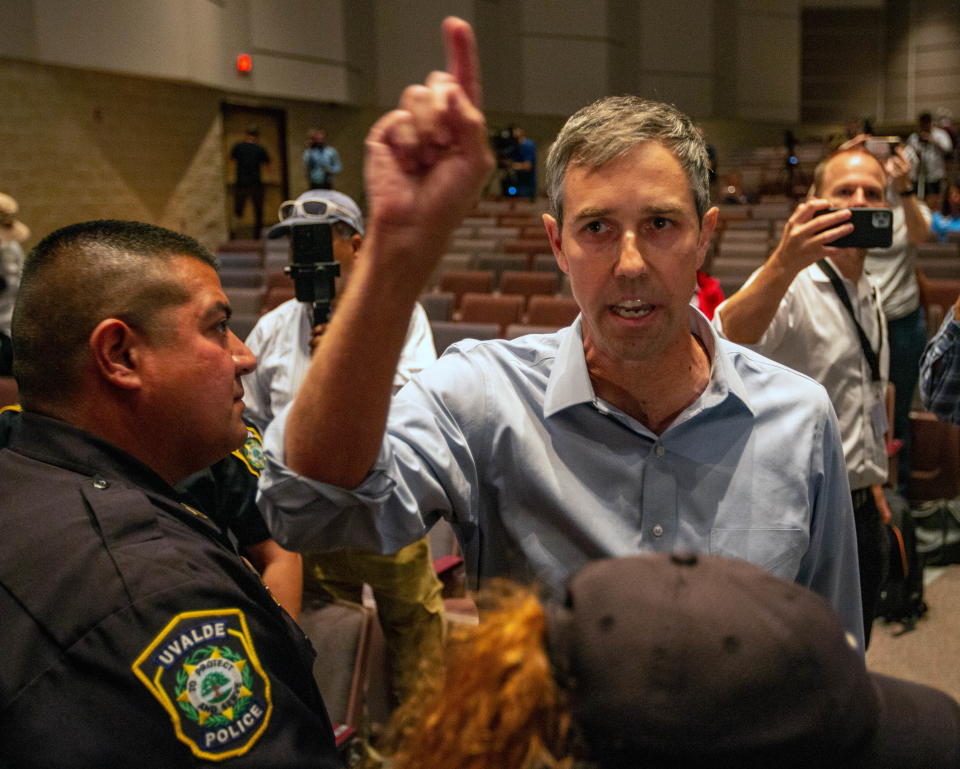 Beto O'Rourke stands in an auditorium next to a police officer who stares intently at him as two people behind him hold out phones as though filming the incident.