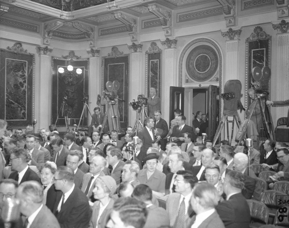 FILE - In this Jan. 19, 1955, file photo photographers have their equipment set up at the back of the room as reporters take their places for President Dwight Eisenhower's news conference in Washington. It was the first conference at which full picture coverage was permitted. Photographer with three cameras at center is John Rooney of The Associated Press. Just below Rooney, Marvin Arrowsmith, AP reporter, enters the room carrying a note board under his arm. (AP Photo/Bill Allen, File)