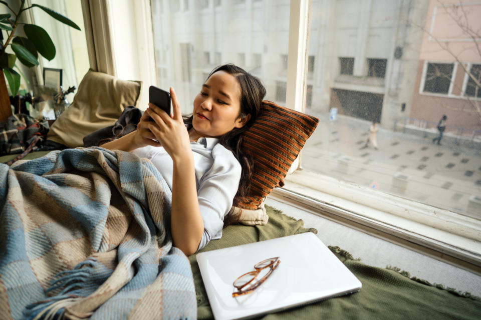 Young asian woman lying by the window on the sofa with a mobile phone in her hands, communicating in social networks. Remote work at home. Online shopping in quarantine period