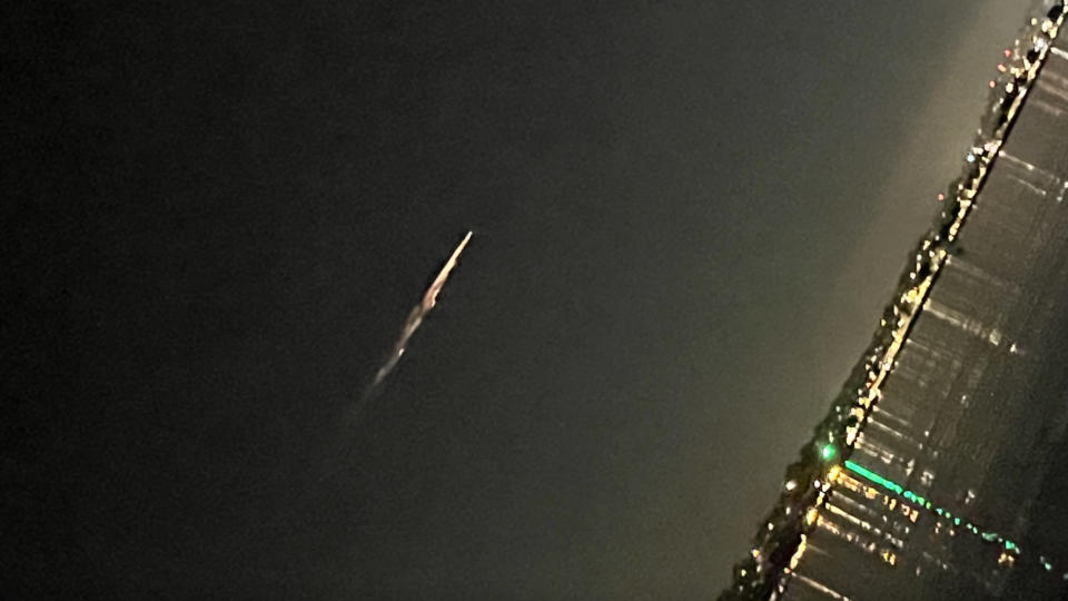 A view of a comet-like streak of light in the sky above water reflecting city lights. The image is tilted such that the horizon is almost vertical on the page, but not quite.